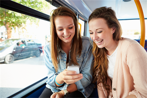 Two smiling teenagers look down at a phone while they ride a bus. 